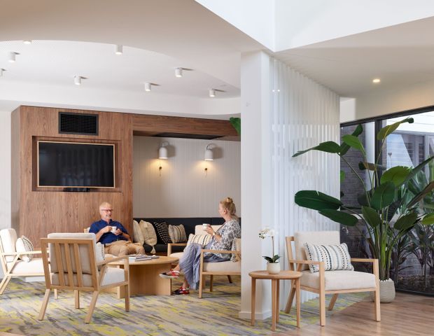 Bright contemporary lounge and reception-style living area with a warm timber entertainment wall, wall-mounted television, built-in seating nook, timber-framed armchairs, patterned cushions, and a large abstract patterned rug. A tall indoor plant sits beside full-height glazing, while white fluted wall panelling and recessed and track lighting add texture and brightness.