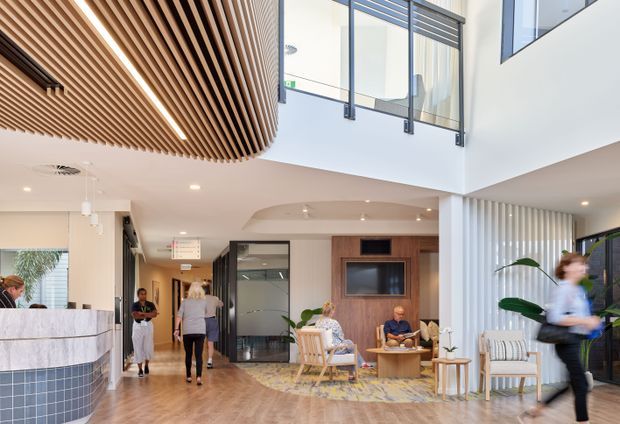 Bright contemporary commercial lobby or reception area with a double-height void, open mezzanine, floor-to-ceiling glazing, and a prominent curved timber slatted ceiling feature. The space includes a stone reception desk, timber flooring, wood-panelled media wall, informal lounge seating, vertical white fins, and several people using the area.