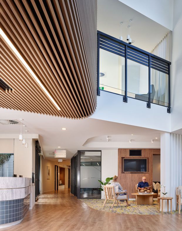 A bright contemporary lobby or reception area with a double-height void. The space features a striking suspended timber slatted ceiling, polished dark-framed internal glazing and balustrades at an upper level, warm timber-look flooring, a curved tiled reception desk, and a timber feature wall with an integrated television. Lounge seating and pendant lights create a welcoming hospitality or commercial atmosphere.