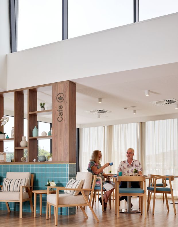 Bright, modern café or hospitality interior with a double-height glazed upper level, warm timber shelving and columns, turquoise tiled counter, light timber flooring, and coordinated café seating. Sheer curtains diffuse abundant daylight, while two people sit at a small dining table. The space has a clean, welcoming, contemporary resort-like feel.