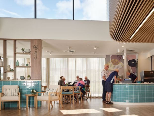 Bright contemporary café or hospitality interior with a turquoise square-tiled service counter, timber flooring, light timber furniture, large sheer-curtained glazing, decorative shelving, and a distinctive slatted timber ceiling feature. Several people are gathered around dining tables and the counter.