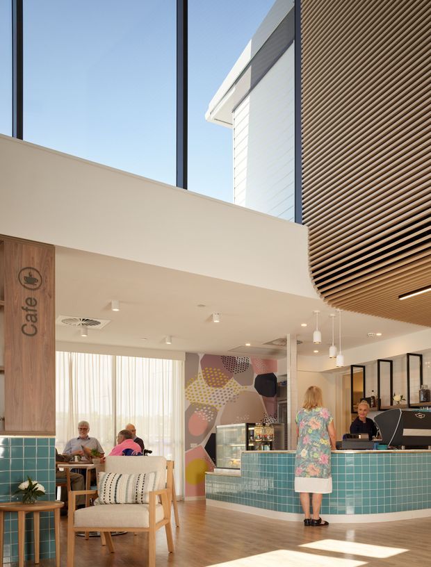 Bright contemporary café interior with a prominent turquoise tiled service counter, timber flooring, light timber seating, pendant lights, and a colourful geometric mural. A timber-clad café sign, large windows with sheer curtains, black-framed glazing, and a slatted timber ceiling feature create a welcoming, airy hospitality setting.