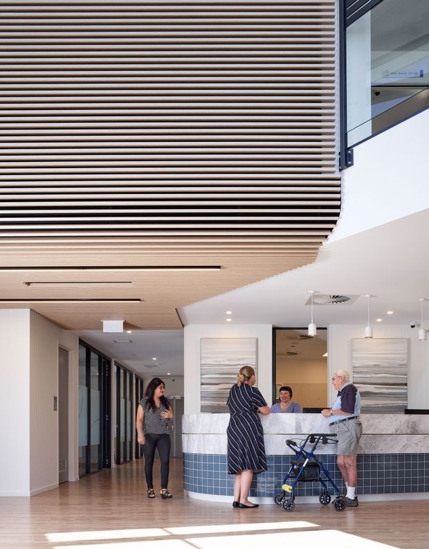 A bright contemporary reception lobby with a curved reception desk clad in white marble-look panels and dark blue square tiles. The space features extensive light timber slatted ceiling and wall detailing, full-height dark-framed glazing along the corridor, pendant lights, recessed lighting, and large abstract coastal-toned artworks. Several people are gathered at the desk, including a person using a walker.