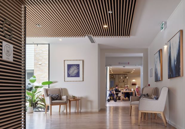 A bright, refined interior corridor or waiting area with warm timber flooring, white walls, framed artwork, upholstered armchairs, and indoor plants. A striking slatted timber ceiling feature extends through the foreground, while a reception or lounge area is visible beyond the opening. The space appears to be a contemporary healthcare or commercial interior, with clear wayfinding and an accessible, welcoming layout.