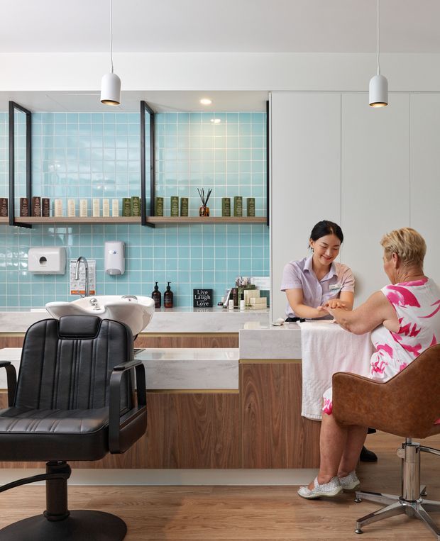 A bright, contemporary hair salon interior with two styling stations, black salon chairs, a white basin, timber cabinetry and benchtops, and a turquoise square-tile backsplash. A stylist is attending to a seated client at the right-hand station. Pendant lights and open shelving with product displays line the wall.