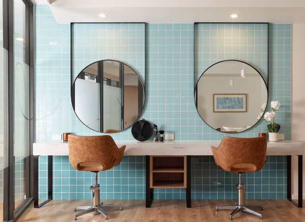 A stylish shared bathroom vanity with two round mirrors, twin wall-mounted basins or countertop vanity positions, and two tan leather swivel stools. The wall behind the vanity is finished in glossy turquoise square tiles with dark grout, while the vanity top is a pale stone or solid-surface slab supported by dark open shelving. Full-height glazing is visible on the left, and warm timber-look flooring, recessed ceiling lights, framed artwork, and small decorative items complete the space.