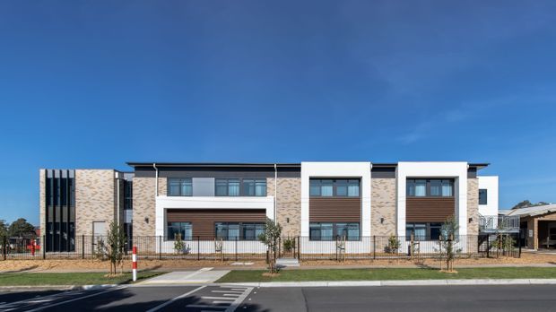 Street-facing exterior view of a contemporary multi-unit residential building with a symmetrical facade, pale textured masonry, dark vertical screening, projecting white-framed window bays, timber-toned horizontal cladding, balconies, and a landscaped setback behind a low metal fence. Clear blue sky and roadway are visible in the foreground.