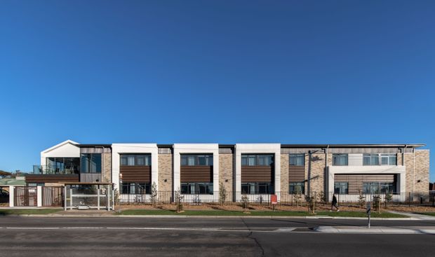 Wide exterior view of a contemporary multi-unit residential building with a long, rectilinear facade. The design combines light masonry or stone-toned wall sections, extensive dark-framed glazing, projecting box-like window bays with horizontal timber-toned screening, and a prominent glazed entry volume. A low metal fence and paved street frontage are visible under a clear blue sky.