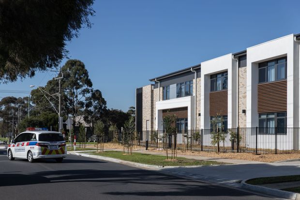 Street-level exterior view of a contemporary multi-unit residential development. The building has a rectilinear form, white rendered sections, light beige brickwork, dark window frames, projecting balconies or recessed terraces, and horizontal timber-look screening. A black metal fence, landscaped verge, driveway, and street frontage are visible.