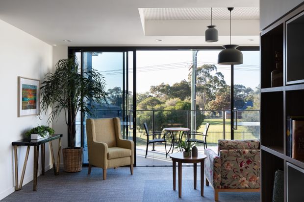 A bright contemporary living room opening onto a balcony or terrace, with floor-to-ceiling black-framed glazing overlooking a leafy suburban landscape. The room features grey carpet, white walls, a woven-texture ceiling recess, black pendant lights, a tall indoor plant, a tan upholstered armchair, floral-patterned lounge chair, small timber side tables, and dark built-in shelving.