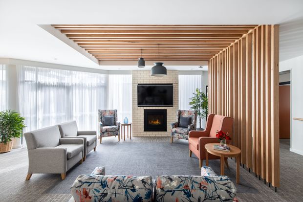 A bright contemporary living room with a neutral grey carpet, large sheer-curtained windows, upholstered armchairs and sofas, and a brick fireplace with a wall-mounted television. A prominent timber slatted ceiling feature extends into a vertical privacy screen, while pendant lights and indoor plants add warmth and detail.