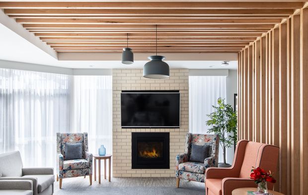 Bright contemporary living room with a central cream brick fireplace and wall-mounted television, framed by large sheer-curtained windows. The ceiling features exposed parallel timber battens, while a vertical timber slat screen defines the right side of the room. Two patterned armchairs, pale seating, pendant lights, and a coral lounge chair create a warm, inviting composition.