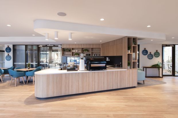 A bright, open-plan hospitality or commercial space centred on a large curved island counter with light timber cabinetry, a white benchtop, and integrated coffee equipment. Behind the counter are open shelves, additional timber joinery, and a dark splashback. The space includes light timber flooring, blue upholstered dining chairs, extensive glazing, and a clean contemporary fit-out.