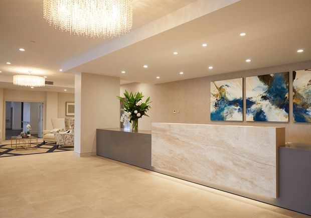 A refined contemporary reception or entry lobby featuring a large stone-clad reception desk with integrated underlighting, warm beige floor and walls, recessed ceiling lights, crystal chandeliers, abstract blue-and-neutral artwork, and a floral arrangement. An adjoining lounge area is visible to the left.