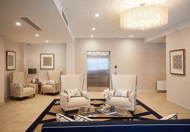 Elegant hotel-style lobby or reception lounge with a central elevator, cream upholstered armchairs and sofas, a glass coffee table, patterned area rug, framed artwork, recessed ceiling lighting, and a large crystal chandelier. The space has warm neutral finishes and a polished, luxurious atmosphere.