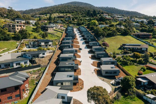 Aerial view of a newly developed residential street on a sloping, heavily landscaped hillside. Compact contemporary homes with flat and low-pitched roofs are arranged in two rows along a central paved access road, surrounded by established suburban houses, gardens, and wooded hills.