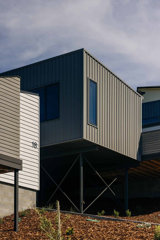 Exterior architectural photograph of a contemporary multi-level building composed of raised and cantilevered volumes. The facade features dark blue-grey and light grey vertical metal cladding, narrow windows, exposed steel bracing beneath the upper level, and a landscaped mulched slope in the foreground. A visible house number '18' appears on the left volume.