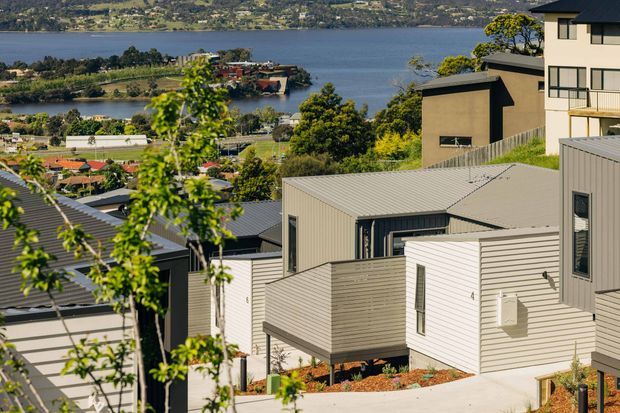 Exterior view of a contemporary multi-level residential development on a sloping site, with several light-coloured homes featuring pitched and low-pitched metal roofs, horizontal weatherboard-style cladding, and dark-framed windows. The neighbourhood overlooks a broad lake and surrounding hills, with landscaped planting and paved outdoor areas in the foreground.