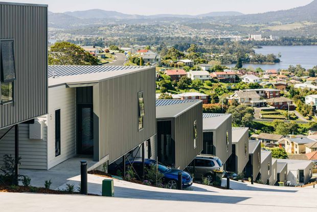 Exterior view of a row of contemporary hillside homes stepping down a steep site, with dark vertical metal cladding, light horizontal siding, and corrugated metal roofs. The elevated homes overlook a dense suburban neighbourhood, a broad body of water, and wooded hills beyond.