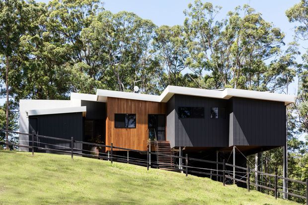 A contemporary single-storey house elevated on a sloping, wooded site. The building has a strongly articulated angular roofline, dark vertical cladding, a warm timber-clad central volume, broad white roof edges, and an open undercroft supported above the hillside. A dark metal fence and steps lead through the landscaped foreground.