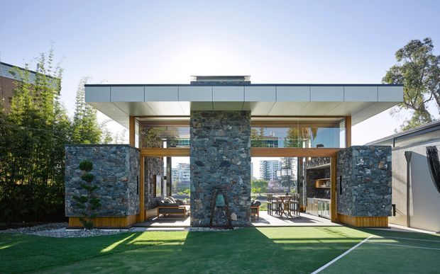 Modern single-storey pavilion-style residence viewed from the rear garden, with broad flat roof overhangs, extensive glazing, warm timber beams and screens, and prominent rough natural-stone walls and central chimney-like pier. The landscaped foreground includes a lawn or synthetic grass court, while living and dining areas are visible through the open glazed facade.