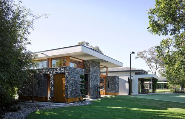Exterior view of a low-profile contemporary residence set within landscaped grounds. The composition shows a flat-roofed main pavilion with extensive glazing, warm timber-framed openings and a dark irregular stone-clad entry volume, connected to additional flat-roofed wings and sheltered outdoor areas. A broad lawn, mature trees and clean modern site lighting surround the building.