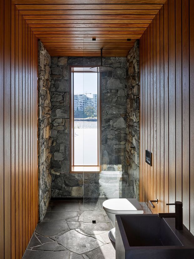 A narrow, dramatic bathroom clad in dark natural stone and richly stained vertical timber panelling. The ceiling continues the timber treatment in a layered framed pattern, while a tall narrow window brings in daylight. A freestanding white bathtub sits beside a dark rectangular vanity with a black basin and matte dark tapware. Irregular stone flooring and walls create a rugged, luxurious retreat-like atmosphere.
