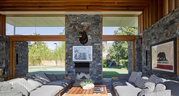 A bright, rustic-contemporary living room with two large grey sofas facing each other across a timber coffee table. Exposed timber beams and warm timber-framed floor-to-ceiling glazing surround substantial rough stone walls, with a central stone fireplace and mounted artwork. The glazing opens views to a landscaped backyard, while patterned cushions and framed art add decorative detail.