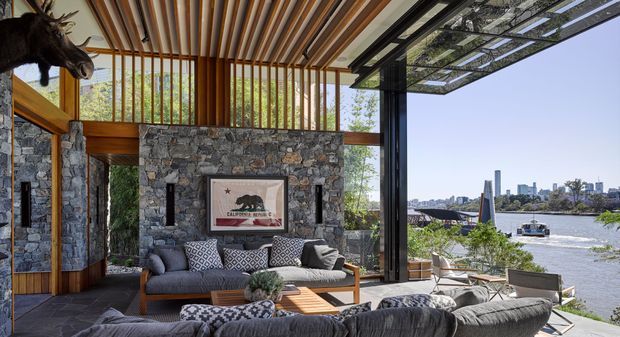 A sophisticated open-plan living room with dramatic floor-to-ceiling glazing overlooking a river and urban skyline. The room features a rugged natural-stone fireplace wall, warm timber framing and vertical battens, a sculptural timber slatted ceiling, dark metal window frames, and layered contemporary seating arranged around a low timber coffee table. A mounted taxidermy-style animal head and framed artwork add a lodge-inspired character.