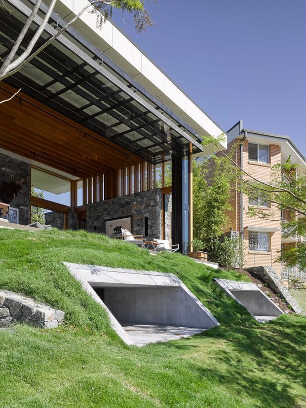 Exterior architectural photograph of a contemporary multi-level residence set into a steep landscaped site. The main structure features a broad flat roof, exposed dark steel framing, extensive glazing, warm timber soffits and vertical timber screens, with substantial dark natural-stone walls. Concrete ramps or recessed access areas cut into the grassy slope in the foreground. A separate brick-clad neighbouring residential volume is visible to the right.