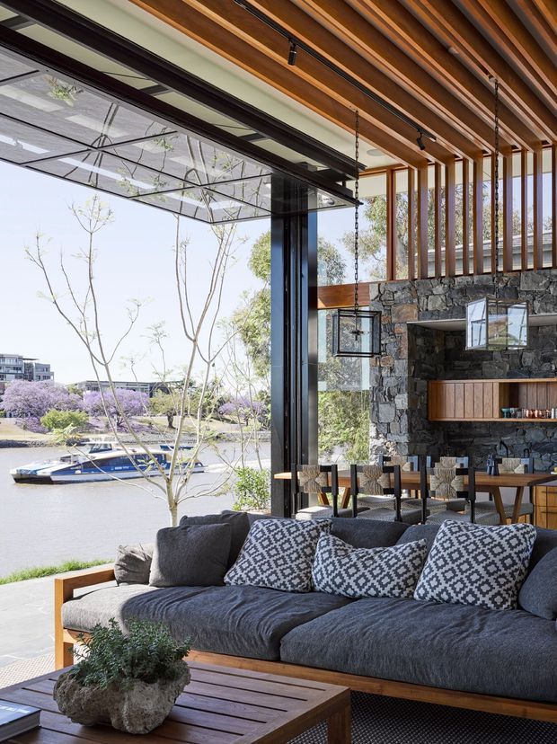 A sophisticated open-plan living and dining space with expansive black-framed glazing overlooking a riverfront setting. The room features a large charcoal sectional sofa, patterned cushions, a timber coffee table, a dining area, a stone-clad feature wall with integrated timber cabinetry and bar storage, and a striking ceiling of exposed warm timber beams and rafters. The indoor-outdoor connection is emphasized by fully opened glazed doors and a covered terrace.