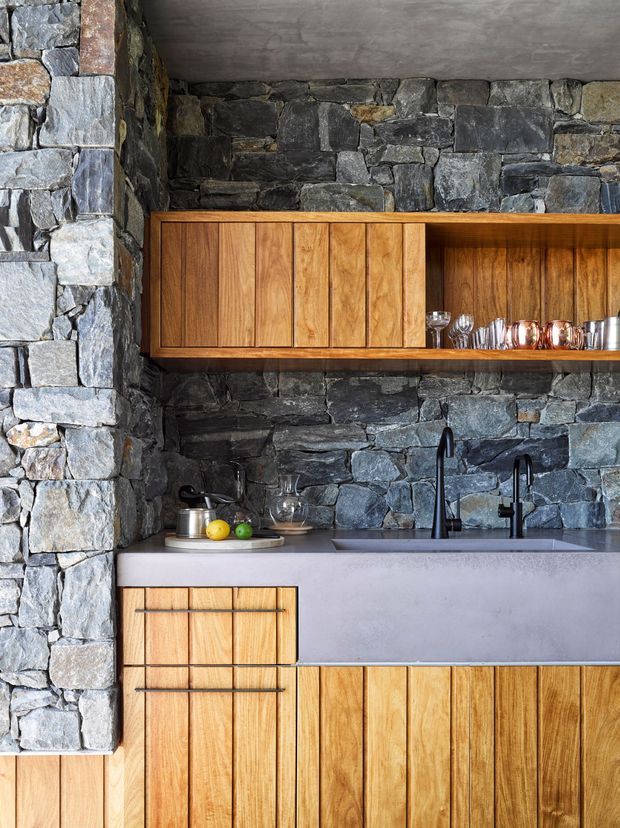Compact rustic kitchen featuring exposed dark grey natural stone walls, warm vertical timber cabinetry, and a pale grey solid-surface benchtop with integrated sink and black tapware. Open shelving displays glassware and copper vessels, creating a tactile, refined mountain-lodge character.
