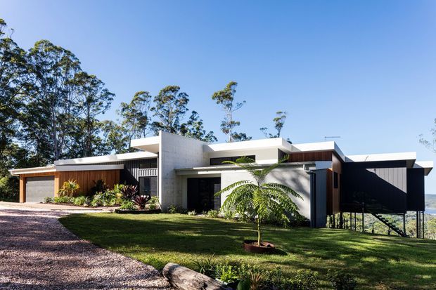 Modern single-storey residence viewed from the front, set on a landscaped sloping site with expansive flat-roofed forms, white rendered or concrete walls, dark timber-toned cladding, and a prominent dark carport or covered entry structure. Large trees and a distant landscape are visible beyond the home.