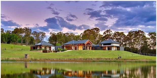 Wide exterior view of a low-rise residential property arranged across a gently sloping landscaped site beside a reflective pond. The buildings have dark hipped and gabled roofs, light rendered walls, timber detailing and extensive glazing, with mature trees and open lawn surrounding the compound under a dramatic evening sky.