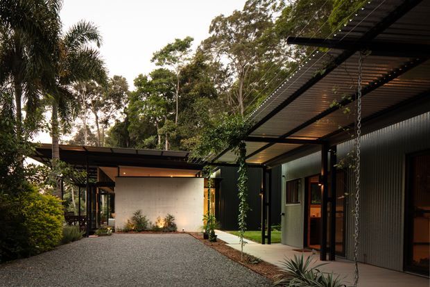 A contemporary single-storey residence set within dense landscaped greenery. The image shows a broad gravel forecourt, low-slung flat-roofed forms, dark slender steel framing, corrugated metal wall cladding, extensive sheltered outdoor circulation, and warm interior lighting visible through glazed openings. Tropical and subtropical planting surrounds the building, while a suspended chain and planted architectural details contribute to an integrated indoor-outdoor character.