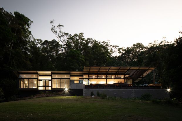 Dusk exterior view of a low-slung contemporary house set within dense bushland. The building features extensive illuminated glazing, a dark horizontal facade, a broad shallow roof with a large covered outdoor terrace or entertaining area, and a low dark masonry retaining wall in front. A lawn occupies the foreground.