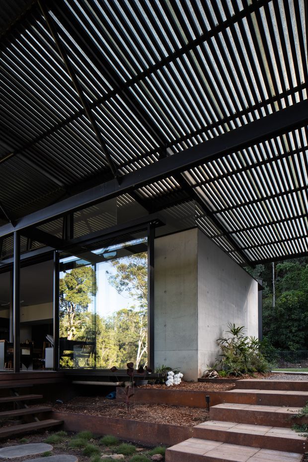 Exterior architectural view of a contemporary house set within landscaped gardens. The image focuses on a dramatic slatted overhead roof or pergola structure with dark metal framing, extensive glazing, exposed concrete walls, and stepped outdoor paths and terraces. Dense vegetation is visible around the building.