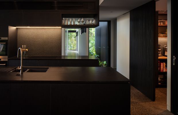 A dark, sophisticated contemporary kitchen viewed from the island, with extensive near-black cabinetry, a large island with integrated sink and tap, a warm metallic textured splashback, and concealed warm LED lighting. A glass-fronted overhead drinks cabinet, open shelving, and a vertically slatted dark partition are visible. The kitchen opens toward a bright glazed passage or courtyard, creating contrast between the moody interior and greenery beyond.