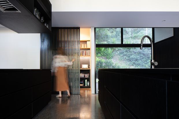 A dark, sophisticated contemporary kitchen viewed toward a large black-framed window overlooking dense greenery. The room features a substantial black island with a tall curved chrome tap, black built-in cabinetry, polished concrete flooring, and a vertical slatted screen separating the kitchen from open shelving and a passage. A blurred person stands near the shelving, while integrated ceiling ventilation and restrained lighting reinforce the minimalist architectural character.