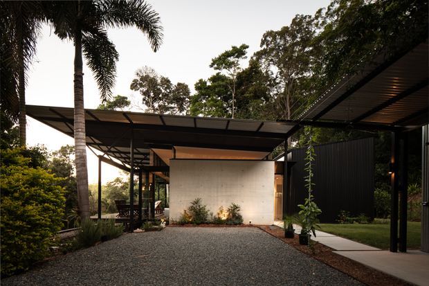 Low, linear contemporary house set within lush tropical landscaping. The view focuses on a broad gravel forecourt and a series of lightweight, flat-roofed steel-framed covered structures, with a raw concrete feature wall, dark vertical cladding, extensive glazing, and a narrow paved walkway leading toward the entrance.
