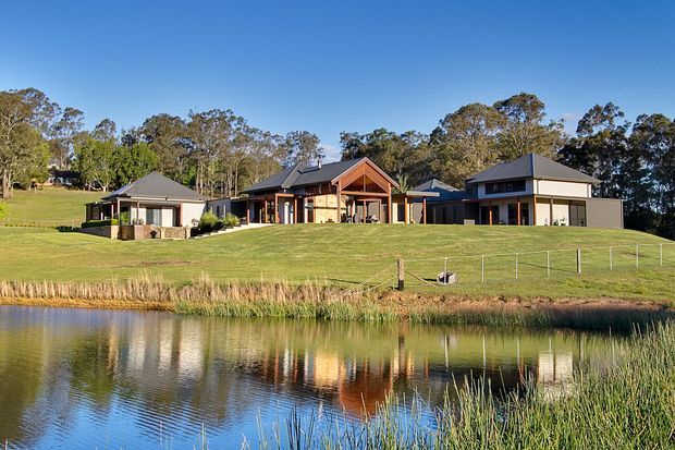 A wide exterior view of a contemporary rural residential property arranged around a central timber-framed pavilion, with two flanking single-storey buildings, dark pitched roofs, pale rendered walls, and extensive glazing. The home sits on a manicured grassy site beside a reflective pond, backed by mature woodland under a clear blue sky.