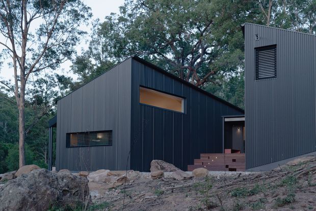 Exterior view of a compact, angular dark-clad house set among mature trees and exposed rocky terrain. The building has a steep asymmetrical pitched roof, corrugated vertical metal cladding, narrow horizontal windows with external louvres, and a raised entry approached by timber steps. The subdued dark facade contrasts with the natural bush setting.