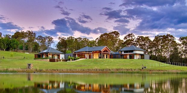 Wide exterior view of a low-rise residential property beside a calm reflective pond, with several connected or closely grouped buildings featuring pitched roofs, timber-toned walls, verandas, and broad glazing. The landscaped grassy site is backed by mature trees and photographed at dusk under a dramatic purple-blue sky.