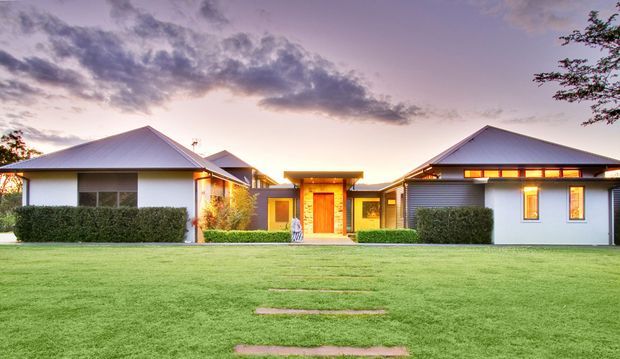 Single-storey contemporary residential home viewed from the front across a broad manicured lawn. The composition features multiple low-pitched hipped roof volumes, light rendered walls, horizontal grey cladding, warm timber-toned entry detailing, landscaped hedges, and illuminated windows and entrance at dusk.