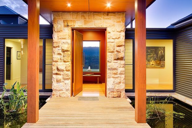 A warmly lit contemporary home entrance viewed from an exterior timber deck. The entry is framed by substantial timber posts and a stone-clad portal, with an open timber door leading toward a window and distant wooded landscape. Dark horizontal cladding, large glazed openings, recessed lighting, and planted water features flank the walkway.