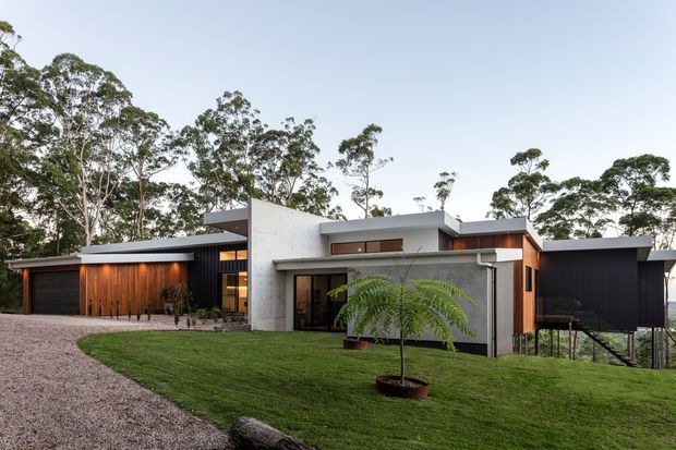 A low, modern residential house set within a wooded landscape. The composition features crisp white rendered or concrete-like wall planes, dark charcoal cladding, warm timber garage and entry detailing, broad horizontal rooflines, and extensive glazing. A gravel driveway and lawn lead toward the house, with mature trees surrounding the site.