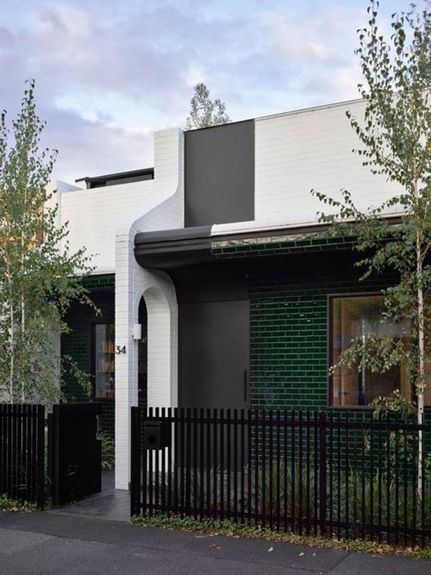 Street-facing view of a contemporary townhouse facade with white horizontal cladding, dark charcoal panels, a curved white entry surround, glossy dark green brickwork, black metal fencing, and a recessed front porch. Trees partially frame the property.
