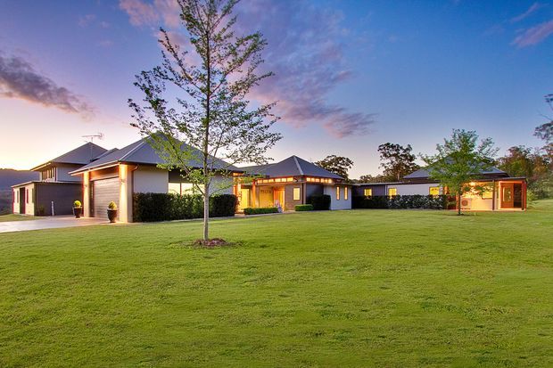 Wide exterior view of a sprawling single-storey contemporary residence at dusk, arranged as several connected pavilion-like wings around a broad manicured lawn. The home has dark pitched roofs, light rendered walls, extensive glazing, warm interior lighting, and timber-toned entry and window details. A rural or suburban landscape is visible beyond the property.