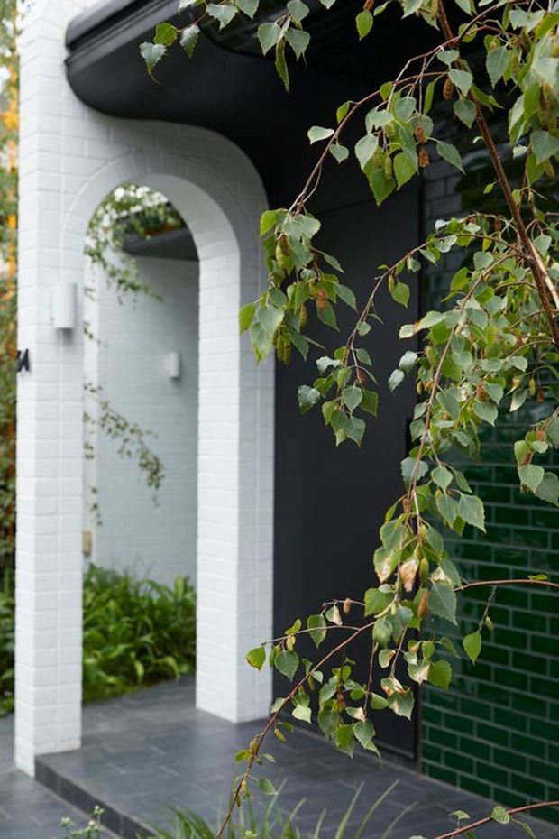 A close-up exterior view of a contemporary residential entryway, featuring a white-painted brick arched portal, dark charcoal facade and soffit, dark tiled paving, and a deep green glazed brick wall. Leafy planting partially frames the entrance and a covered passage beyond.