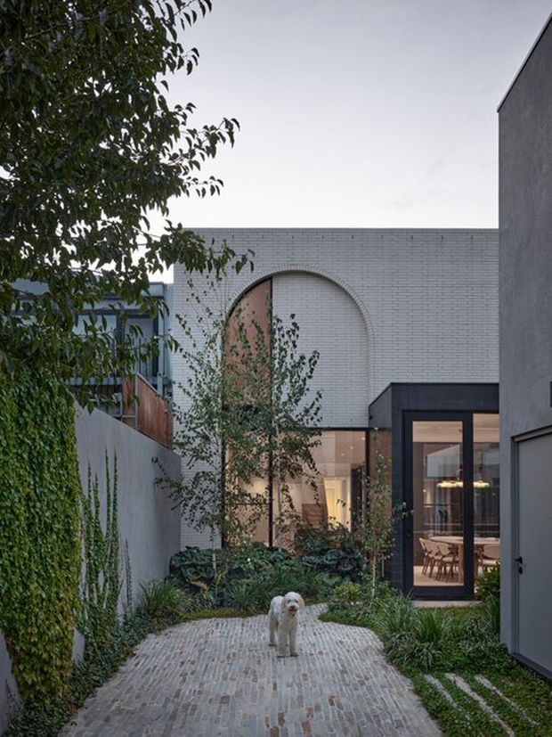 A landscaped courtyard passage leading toward a contemporary residence. The scene features pale grey brick walls, a large arched brick feature surrounding tall glazing, dark framed glass doors, a paved brick pathway, planted garden beds, and a small dog in the foreground. The muted dusk-like light creates a calm, refined atmosphere.
